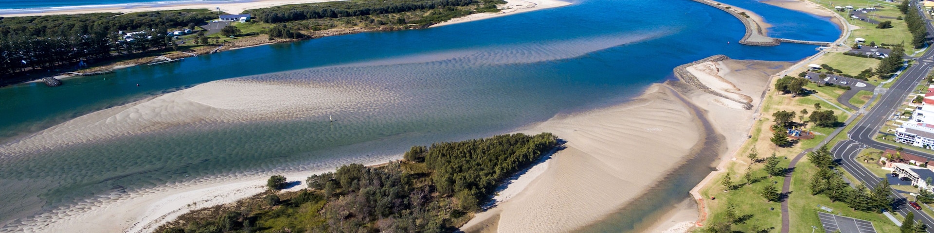 Eine Luftaufnahme einer Küstenlandschaft mit einer weiten Bucht, Sandstränden, einer kleinen Insel mit dichter Vegetation und einer Straße, die zu einer Siedlung führt.