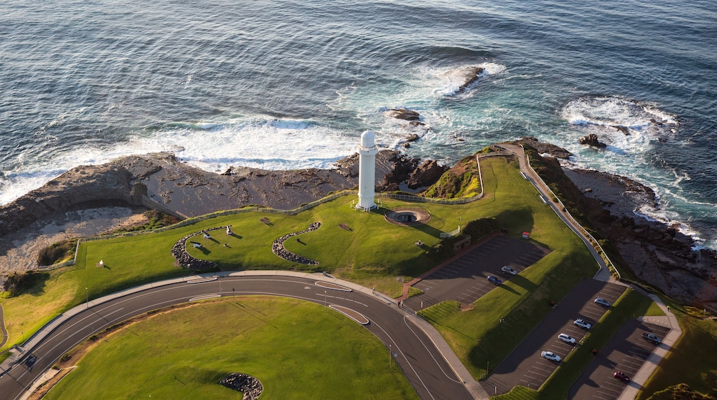 A coastal landscape with a lighthouse, grassy areas, a parking lot, and a road leading to the sea.