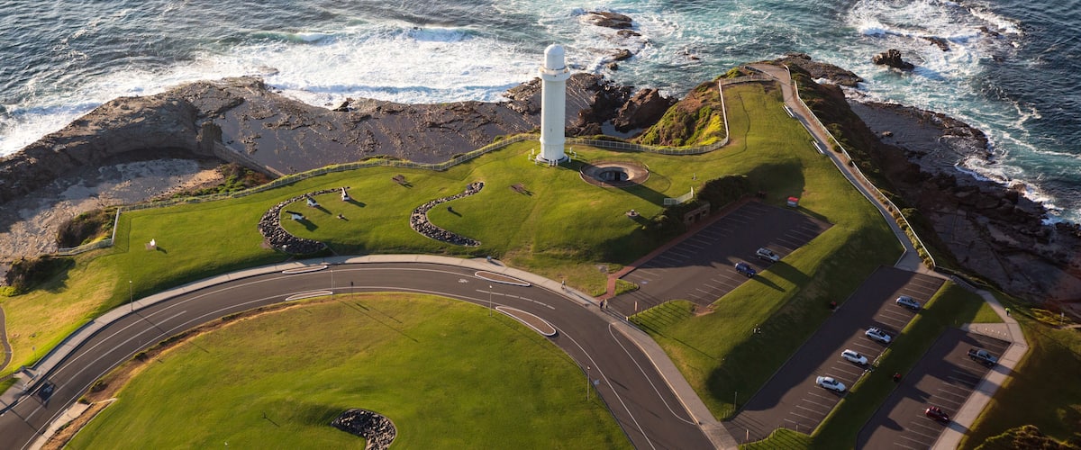 A coastal landscape with a lighthouse, grassy areas, a parking lot, and a road leading to the sea.