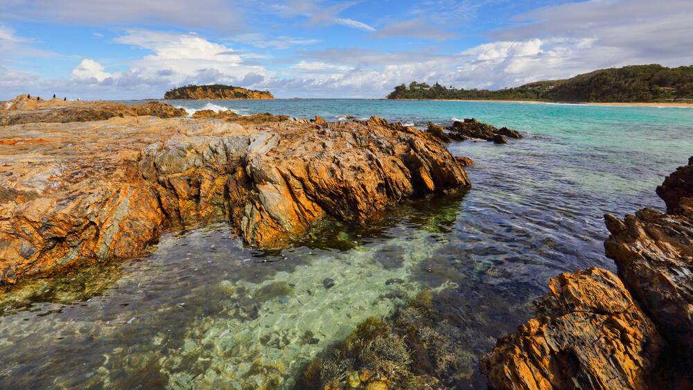 Eine zerklüftete Küstenlinie mit klarem türkisblauem Wasser, eine entfernte Insel und eine hügelige Landschaft.