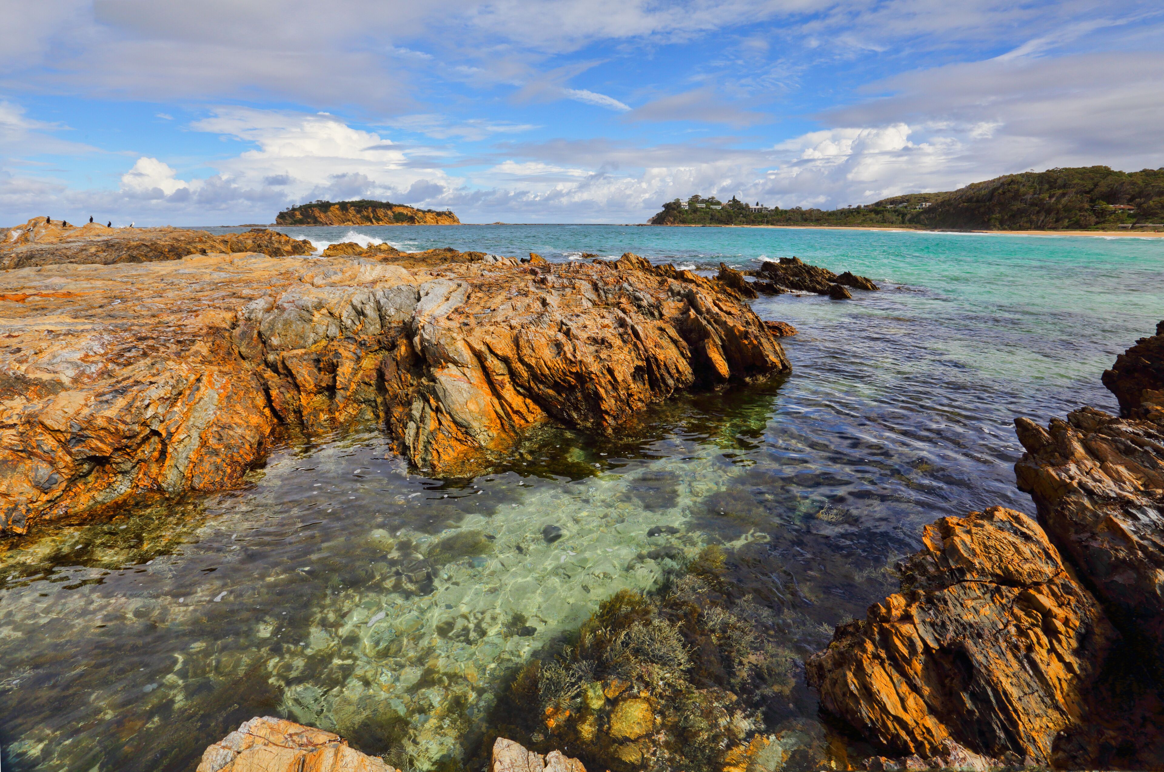 A rocky coastline with clear turquoise water, a distant island, and a hilly landscape.