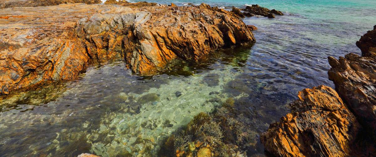 A rocky coastline with clear turquoise water, a distant island, and a hilly landscape.