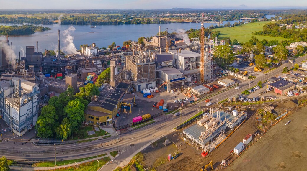 An industrial complex with multiple buildings, smokestacks, and a river in the background.