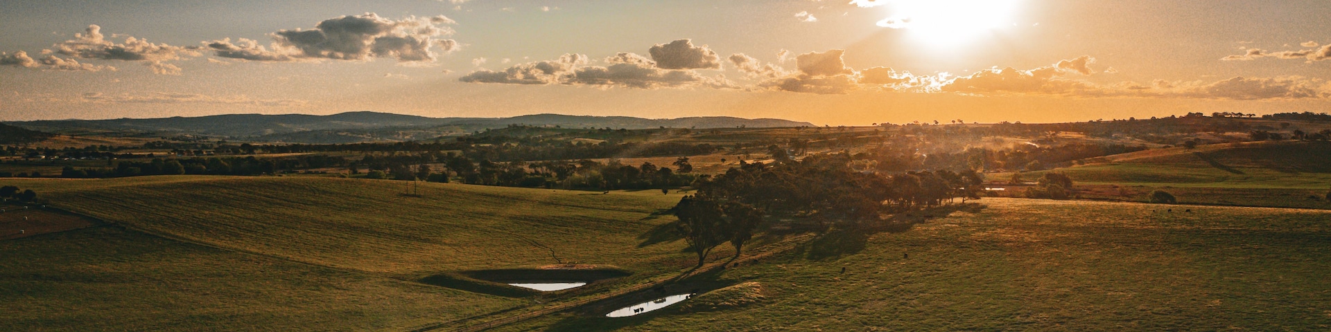 A landscape with a field, a small pond, and distant hills under a partly cloudy sky with the sun setting or rising.