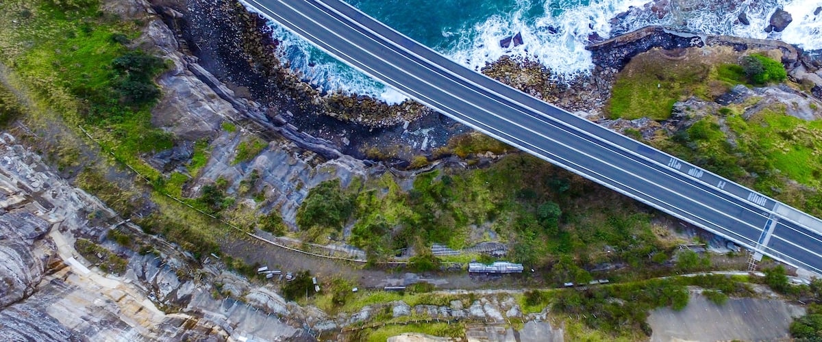 A coastal road with a bridge over the ocean, surrounded by rocky cliffs and greenery.