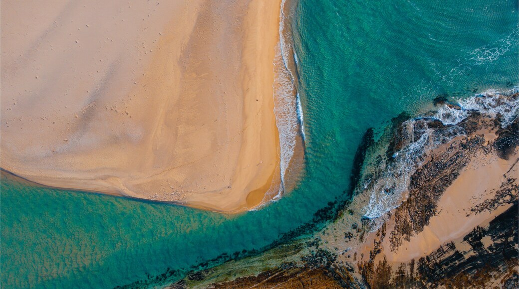 An aerial view of a beach with clear turquoise waters, a sandy shore, and rocky outcrops.