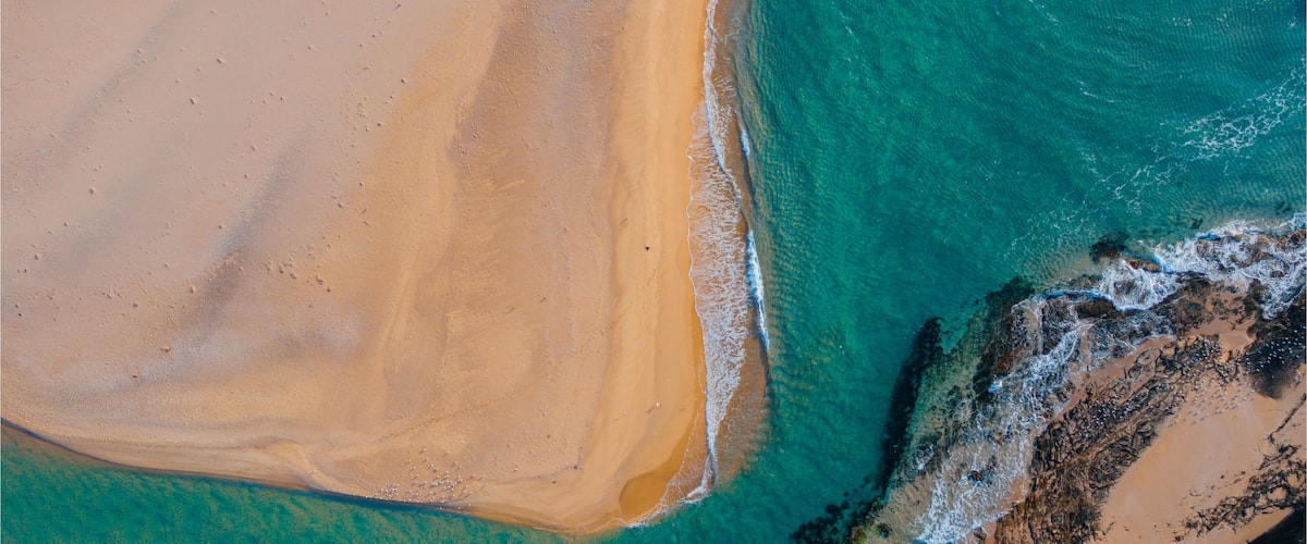 An aerial view of a beach with clear turquoise waters, a sandy shore, and rocky outcrops.
