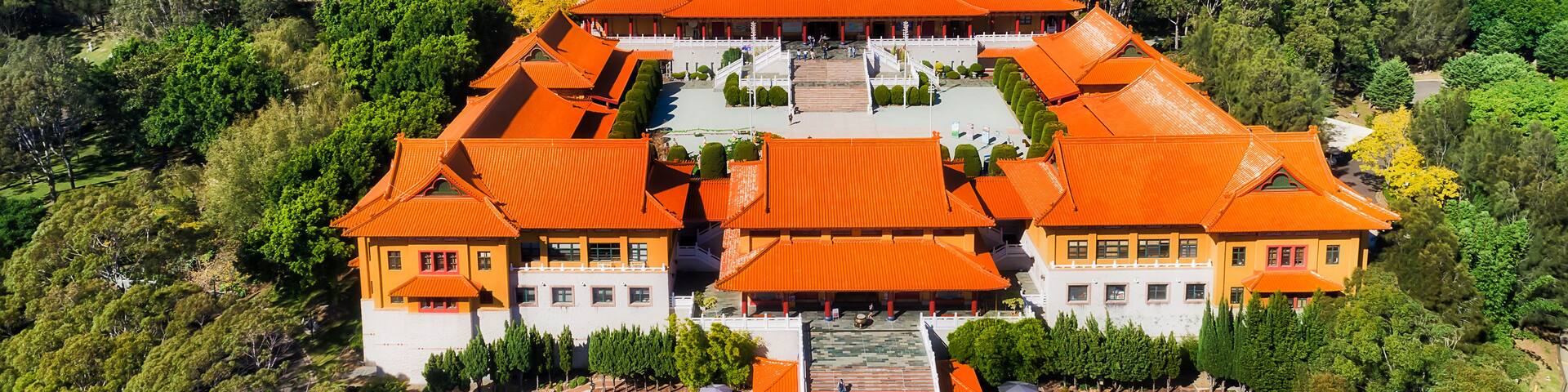A traditional building with a symmetrical roof and multiple wings, surrounded by lush greenery and hills.