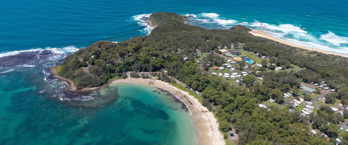 An aerial view of a coastal area with a beach, clear turquoise waters, and dense forested hills.