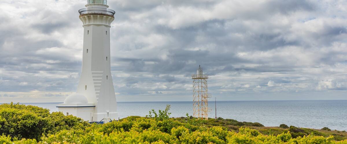 A white lighthouse with a metal tower, surrounded by green vegetation and the sea under a cloudy sky.
