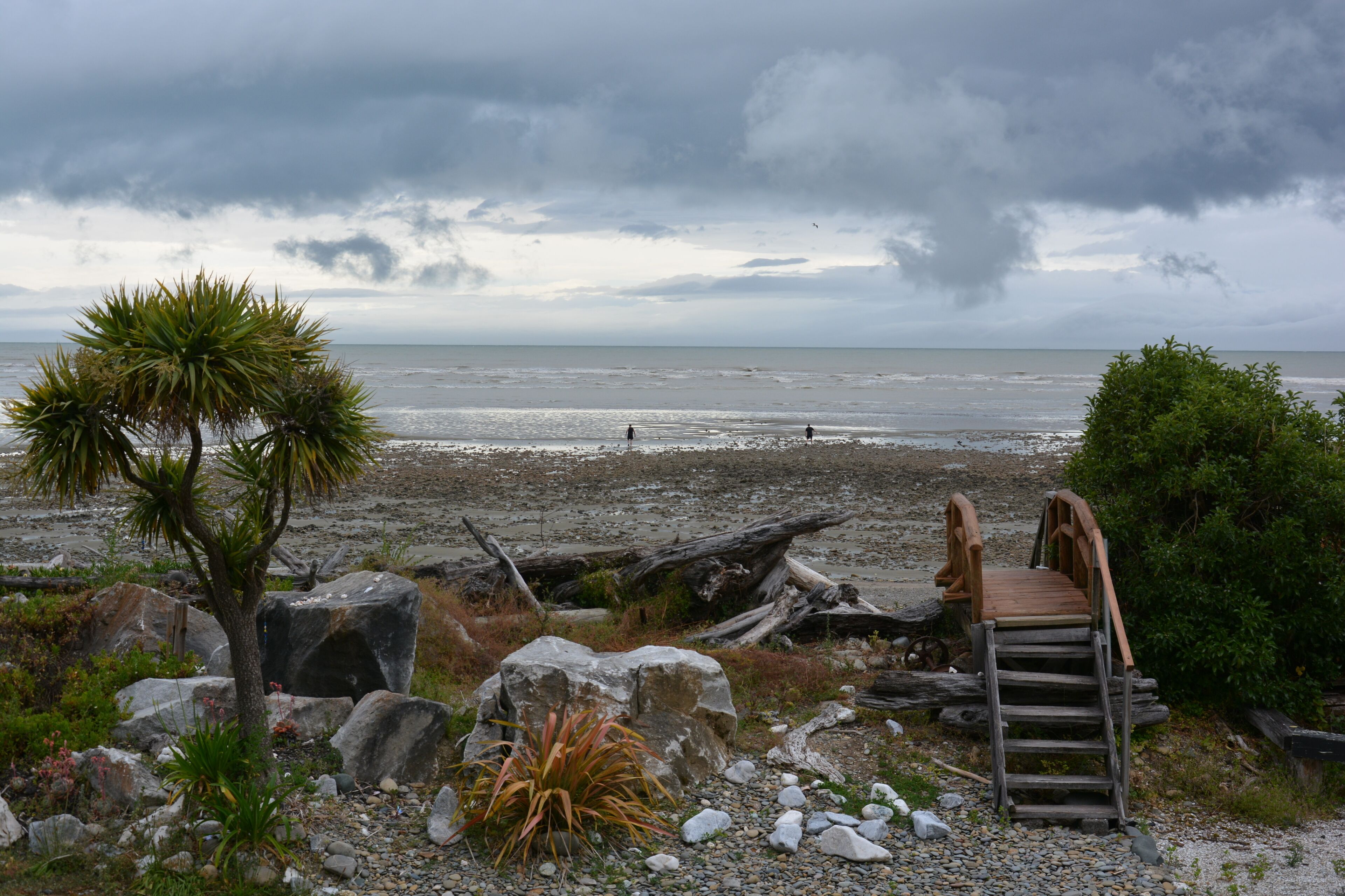 A beach with a wooden staircase leading to a wooden bridge over the water, surrounded by rocks and vegetation.