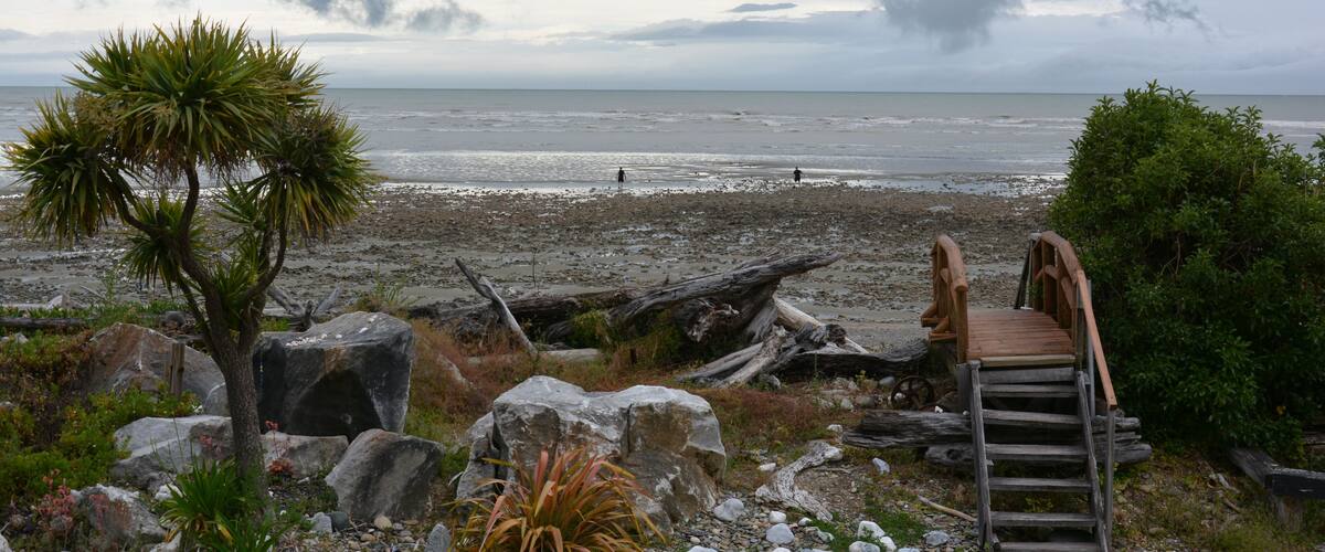 A beach with a wooden staircase leading to a wooden bridge over the water, surrounded by rocks and vegetation.