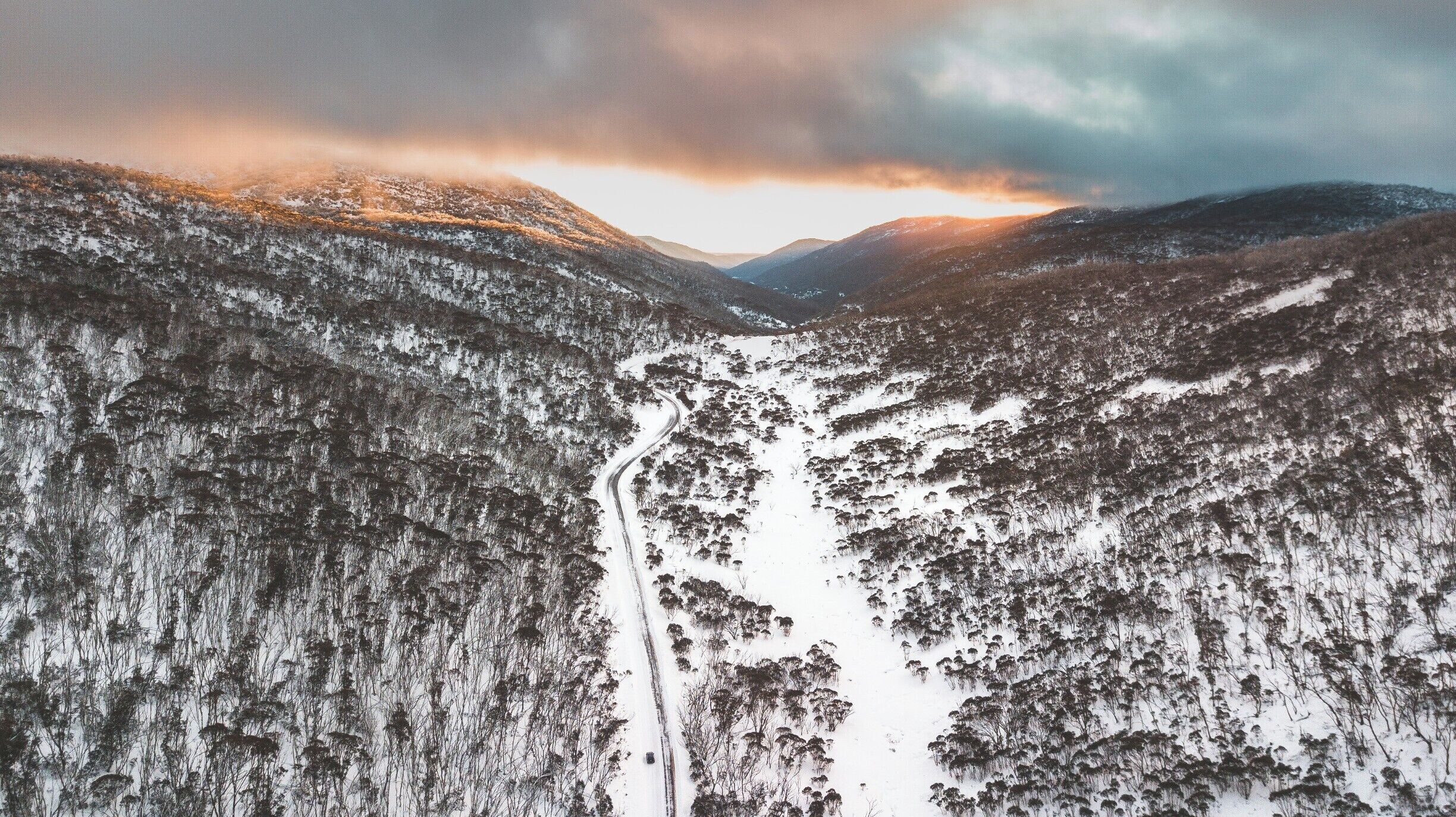 A snow-covered valley with a winding road between forested hills.
