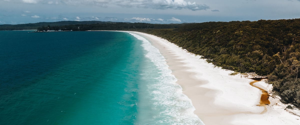 A coastal landscape with a white sandy beach, turquoise waters, and a forested hill in the background.