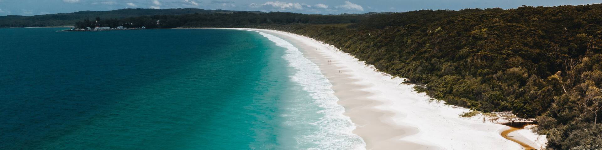 A coastal landscape with a white sandy beach, turquoise waters, and a forested hill in the background.