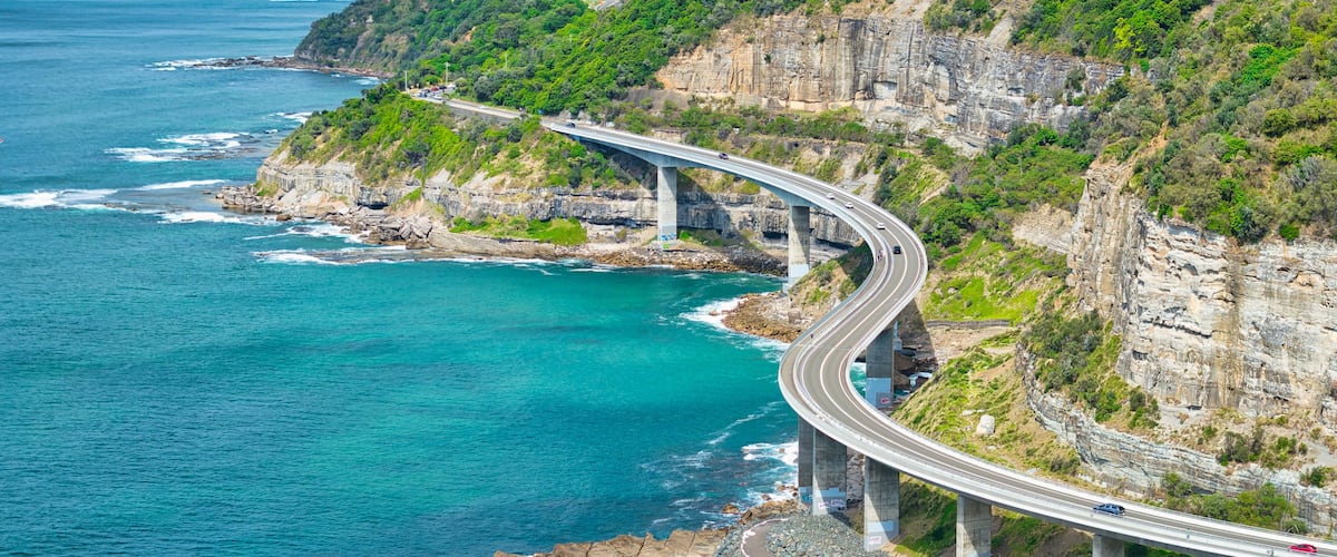 A coastal highway curving along a cliffside with a view of the ocean and distant mountains.
