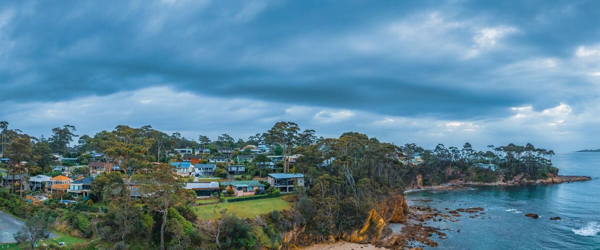 A coastal residential area with houses, dense trees, and rocky shoreline.