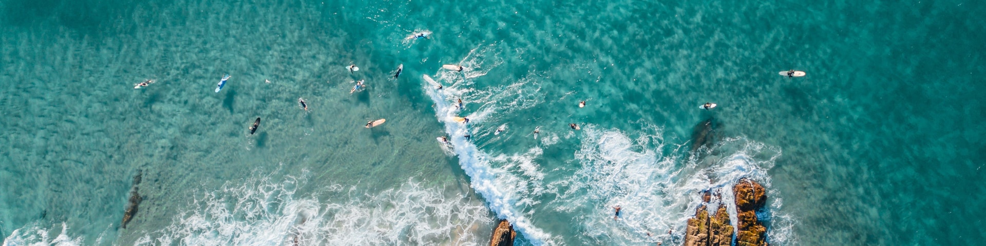 Vista aérea de uma área costeira com águas turquesa cristalinas, falésias rochosas e surfistas na praia.