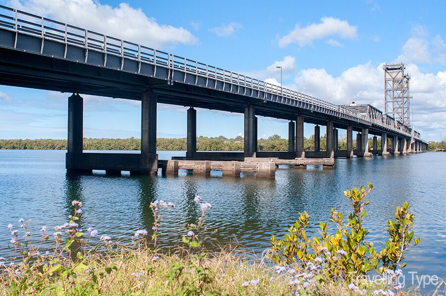A long bridge over a calm river with greenery in the foreground.