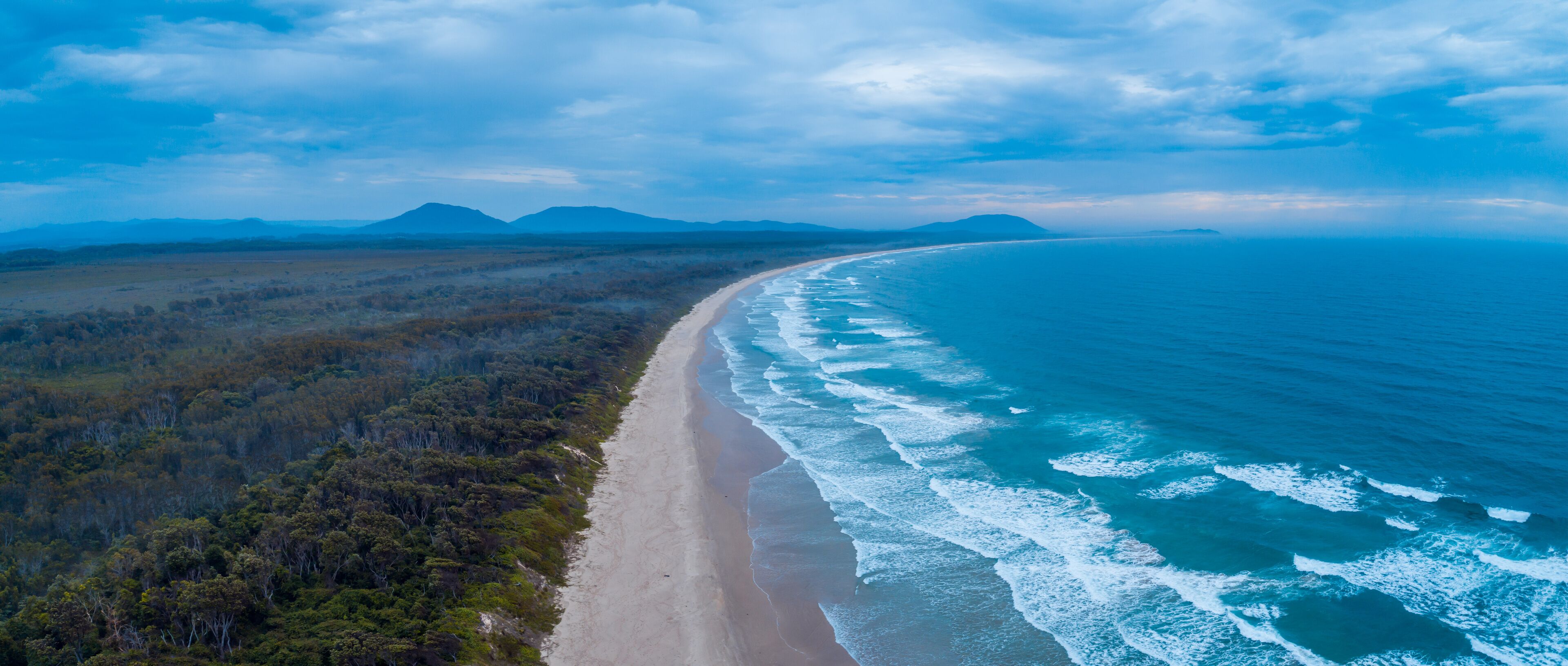A coastal landscape with a sandy beach, ocean waves, and a forested area with mountains in the background.