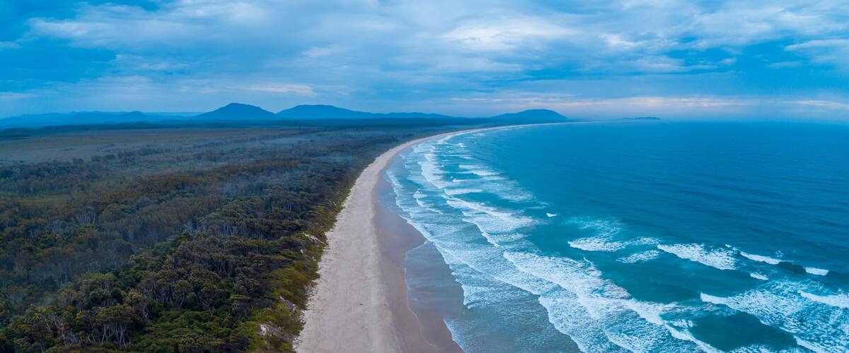 A coastal landscape with a sandy beach, ocean waves, and a forested area with mountains in the background.