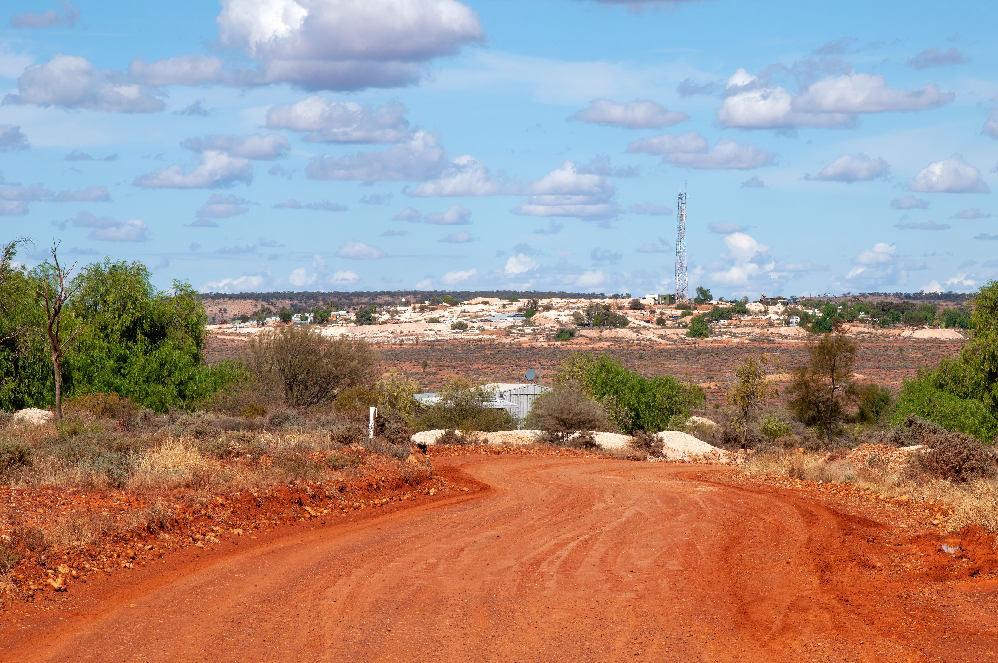 Un camino de tierra serpentea por un paisaje seco y rojizo, con vegetación escasa, un cielo azul despejado y una torre de comunicaciones a lo lejos.