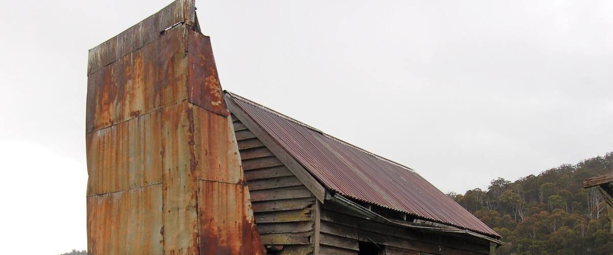 An old, rusted wooden structure with a corrugated metal roof, surrounded by a grassy field and a forested hill in the background.