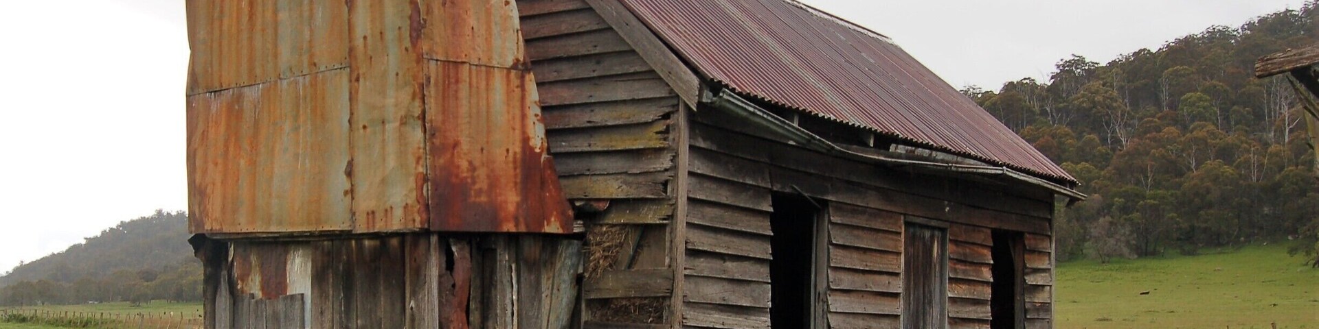 An old, rusted wooden structure with a corrugated metal roof, surrounded by a grassy field and a forested hill in the background.