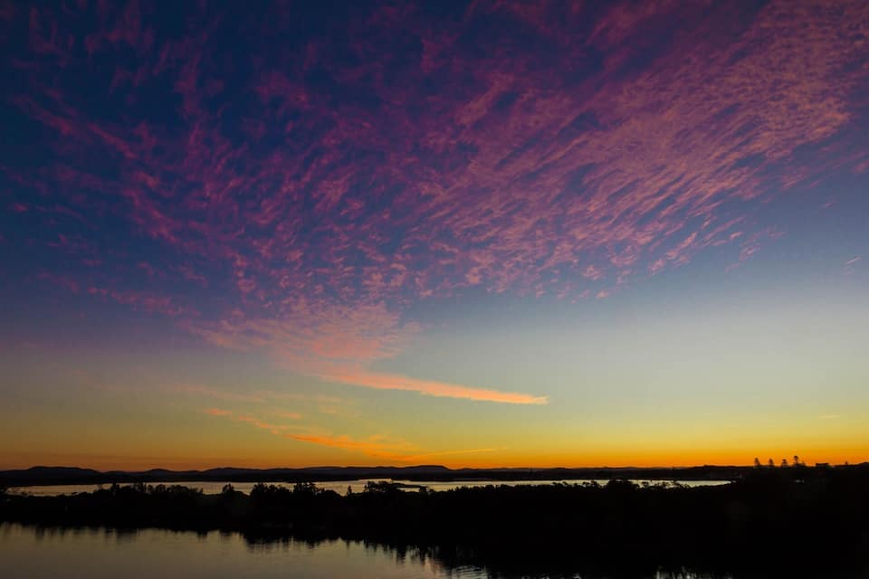 A sunset over a body of water with a silhouette of trees and hills in the background.
