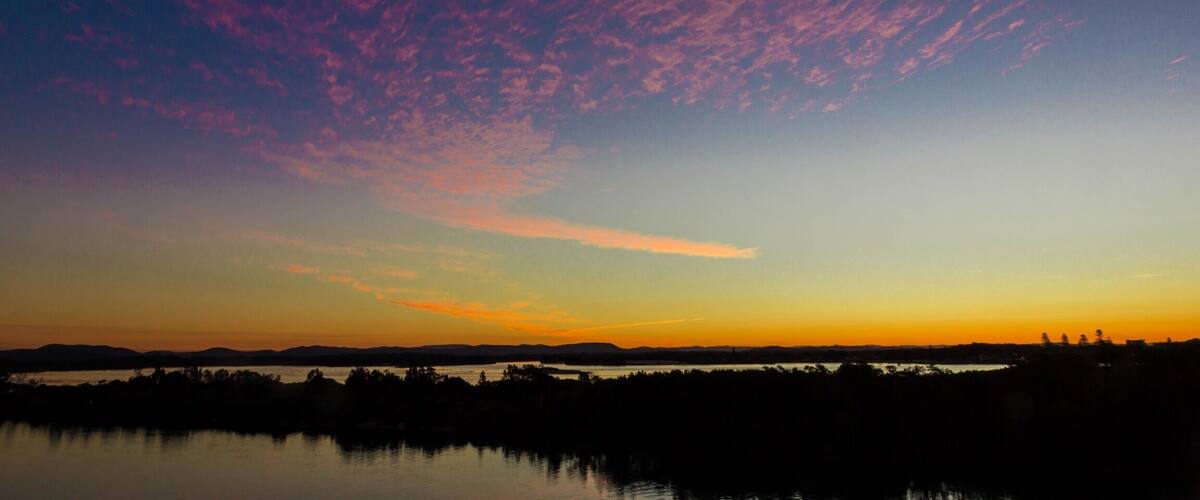 A sunset over a body of water with a silhouette of trees and hills in the background.