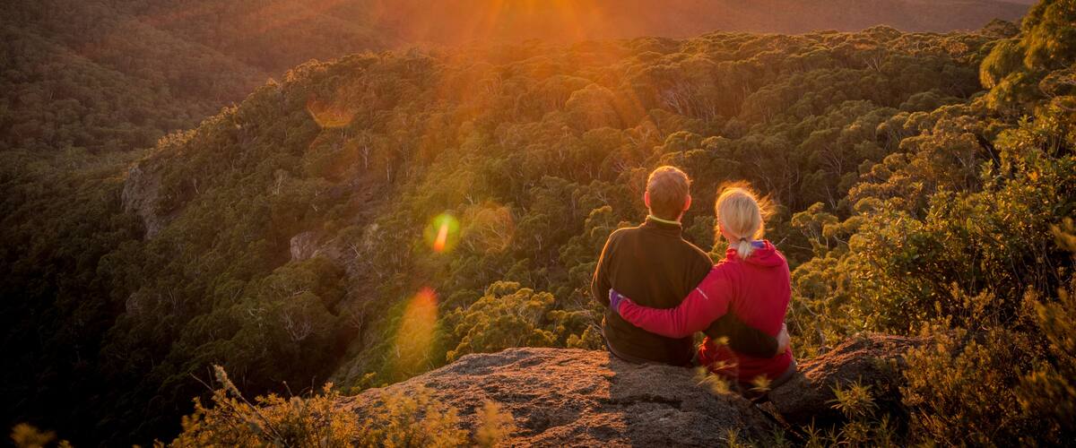 A couple sitting on a rock overlooking a forested valley at sunset.