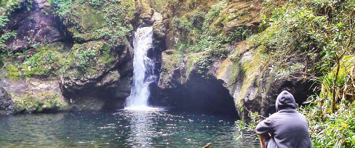 A waterfall in a forested area with a person sitting on a rock by the water.