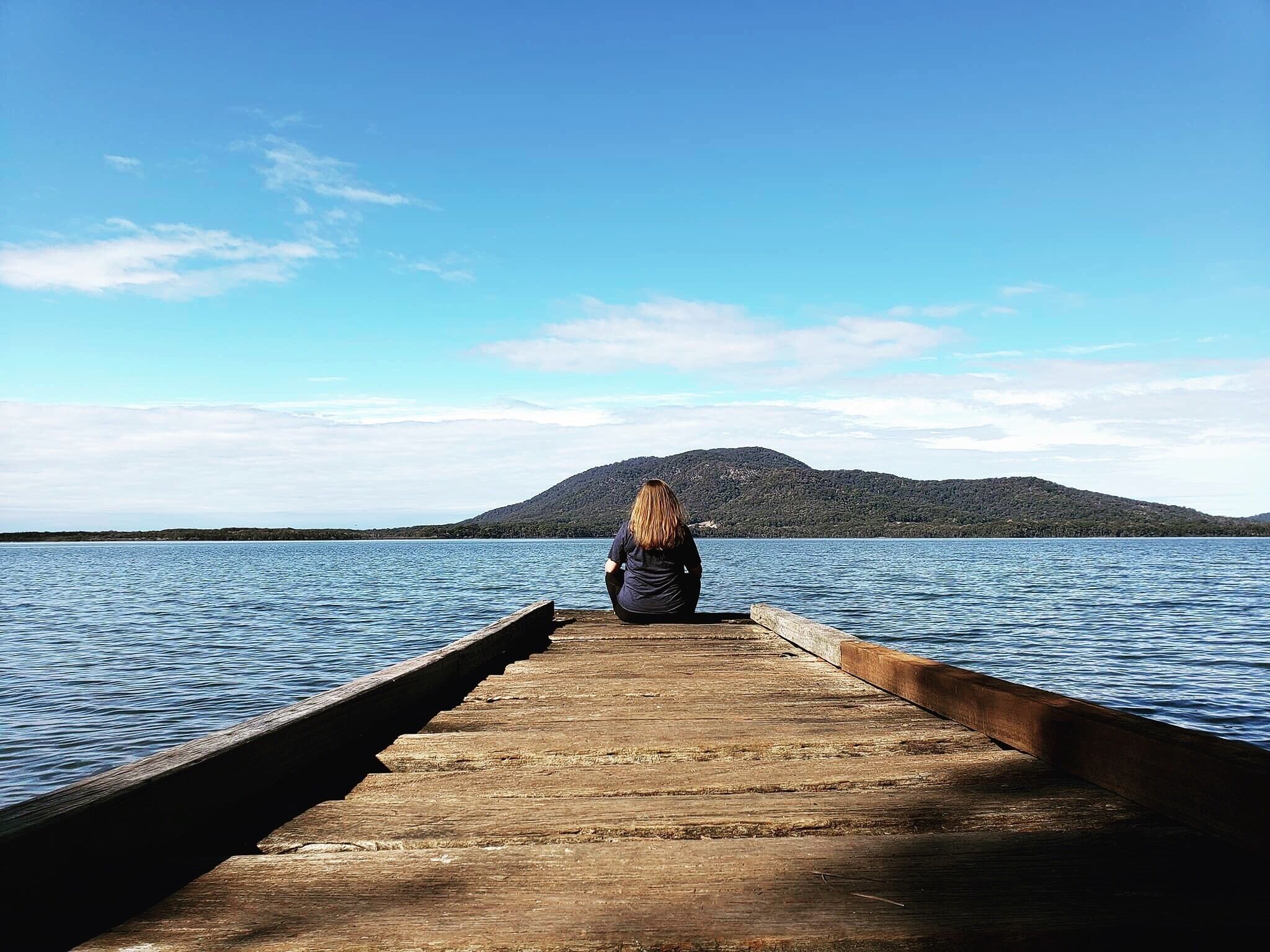 A wooden pier extending into calm water with a person sitting at the end, overlooking a hilly landscape.