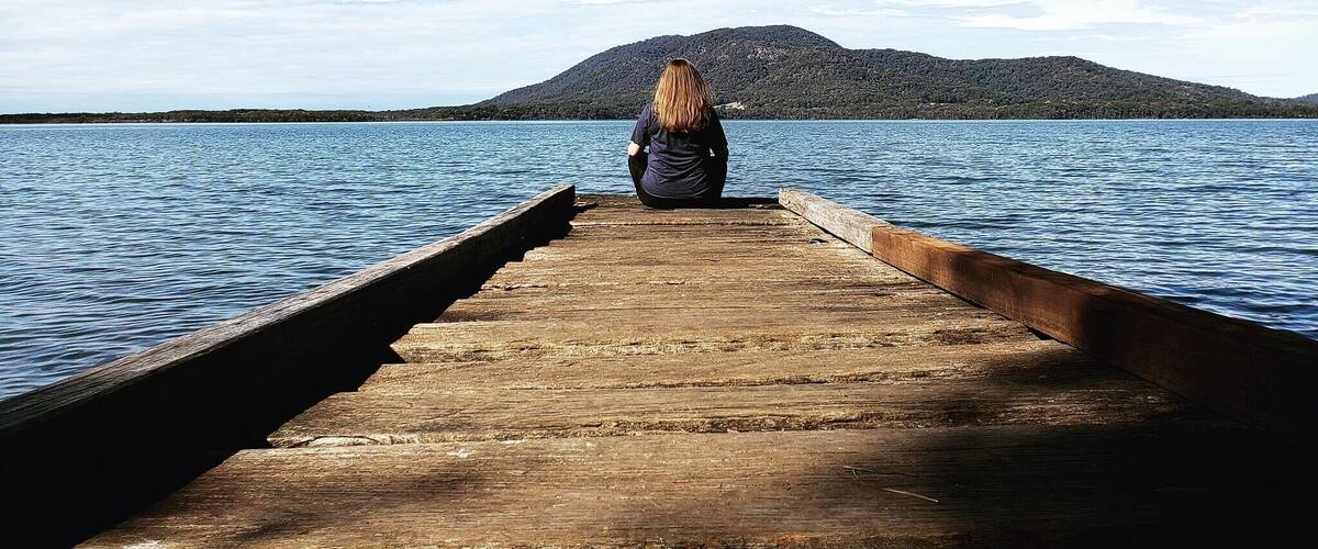A wooden pier extending into calm water with a person sitting at the end, overlooking a hilly landscape.
