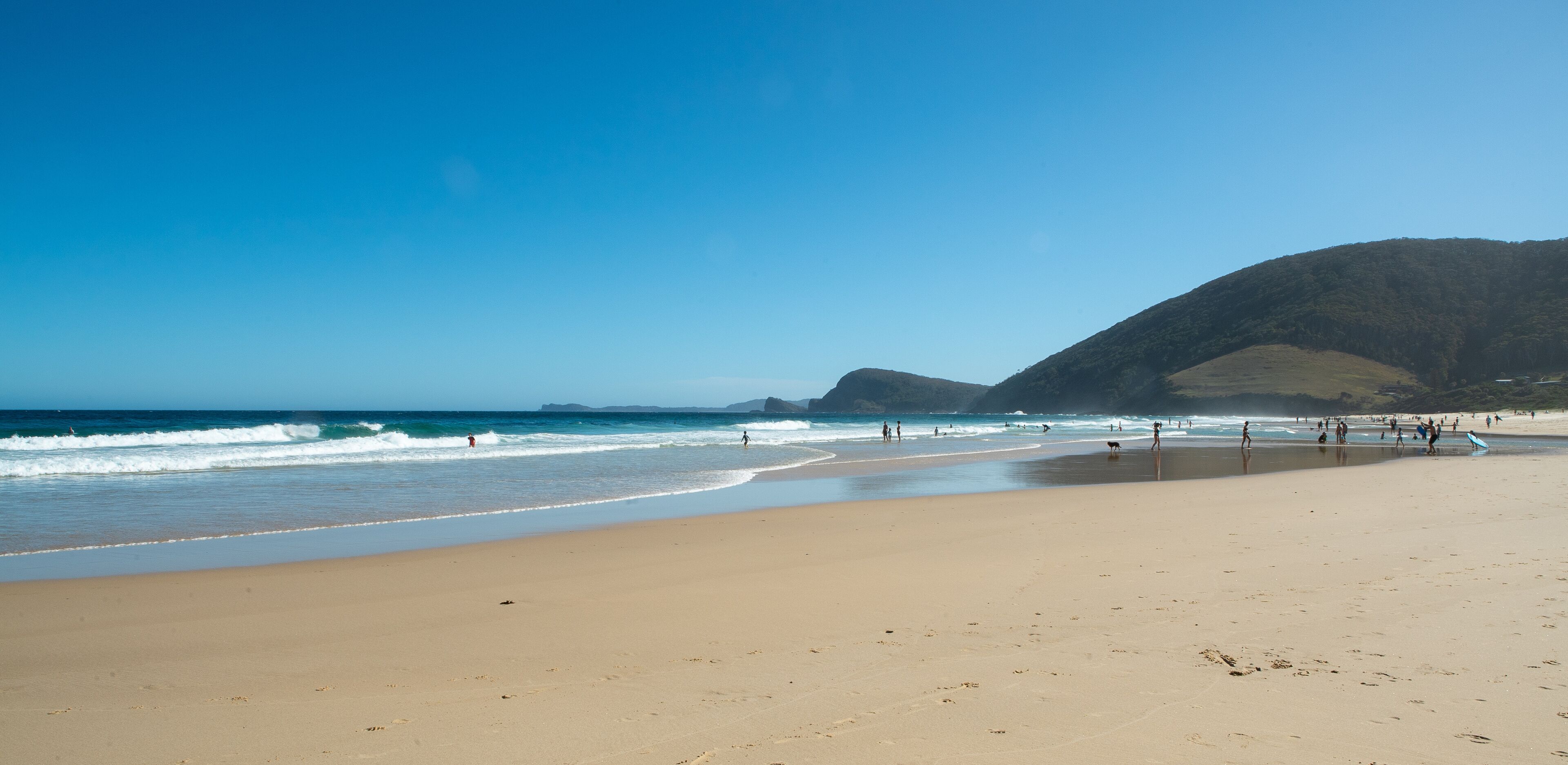 A sandy beach with waves, people, and a mountain in the background.