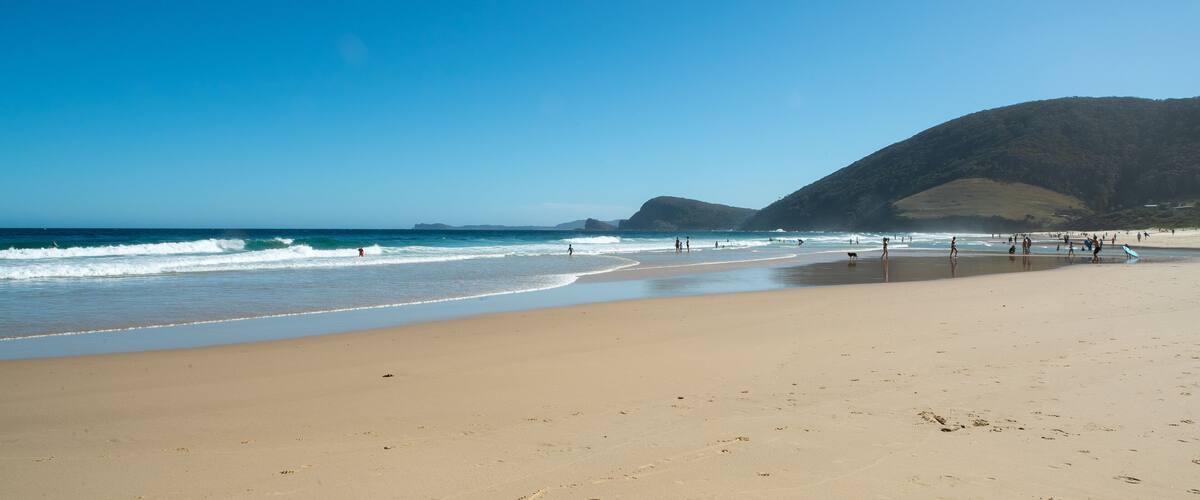 A sandy beach with waves, people, and a mountain in the background.