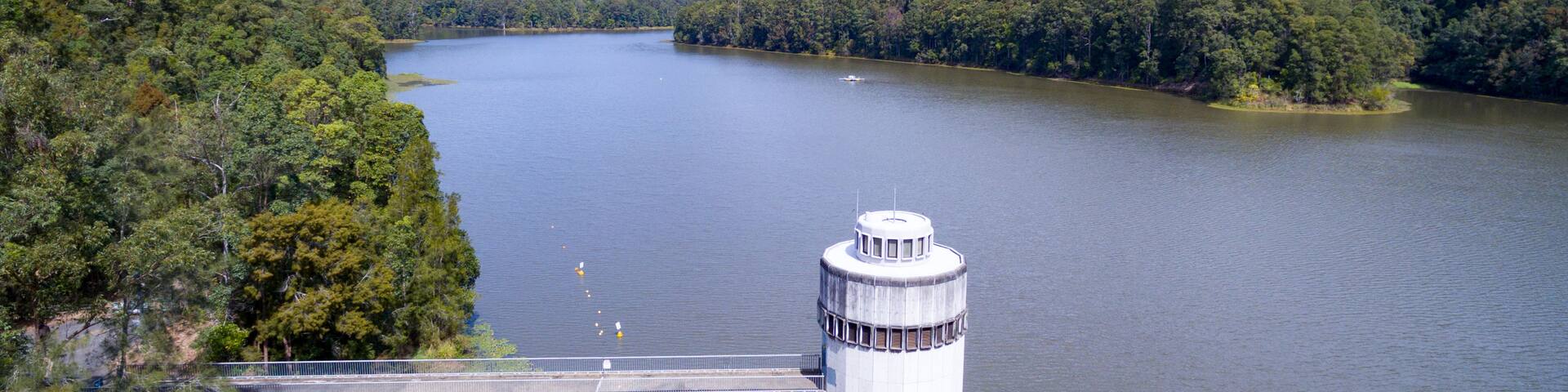A dam with a control tower, surrounded by forested hills and a calm lake.