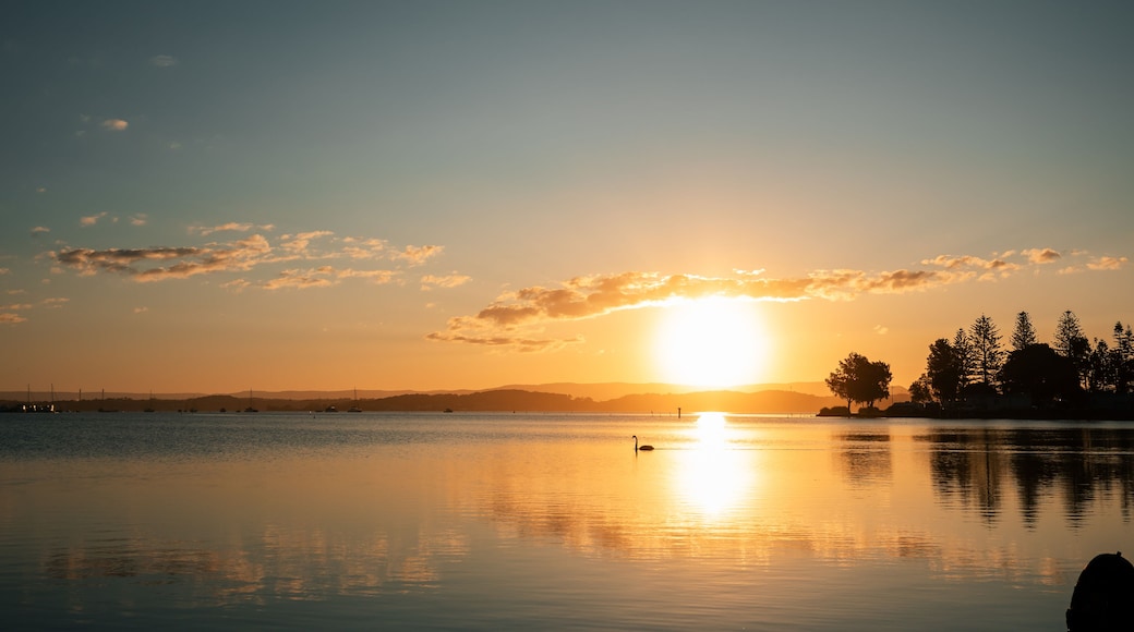 A sunset over a calm lake with boats, trees, and distant hills.