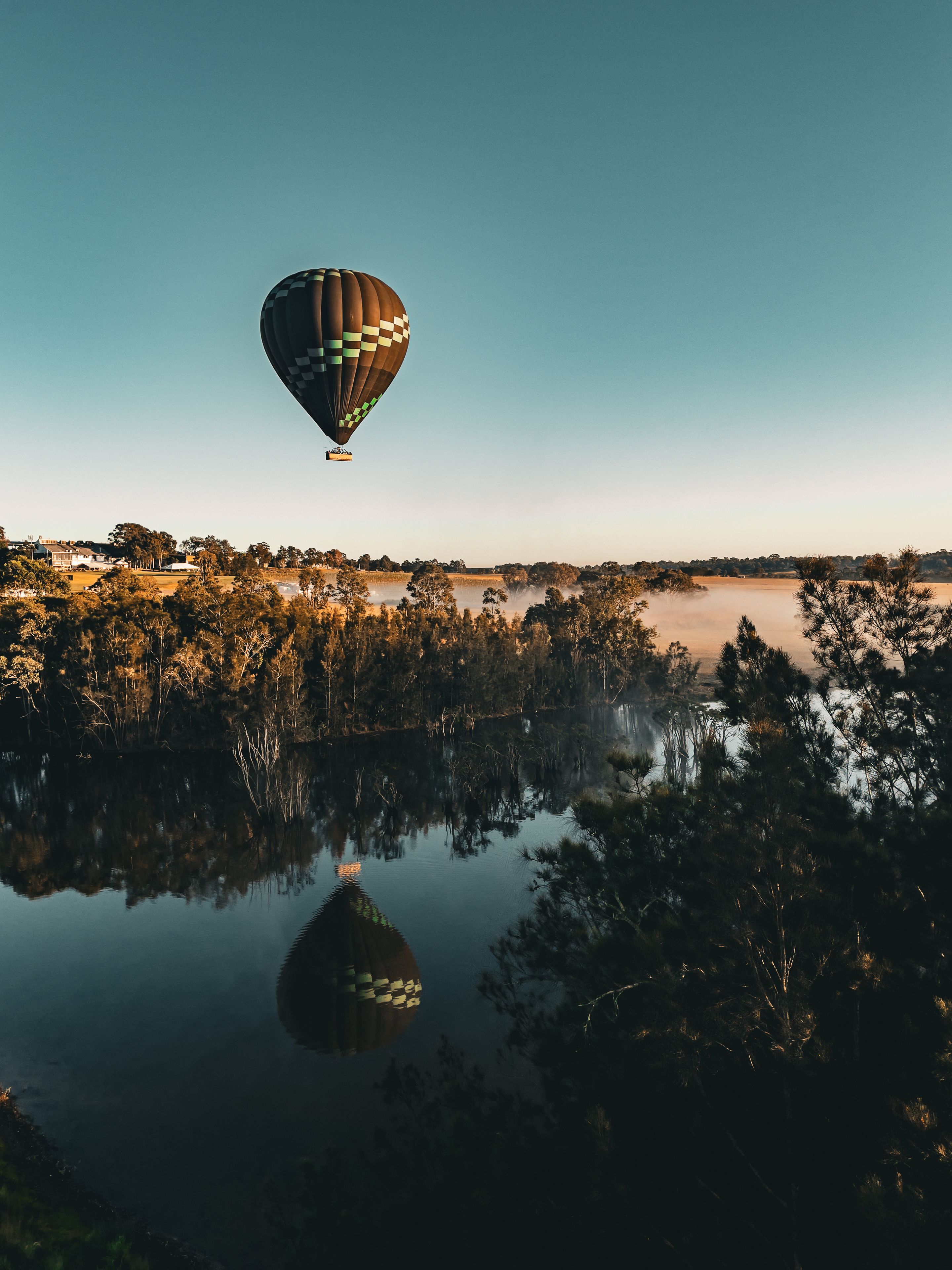 Um balão de ar quente voando sobre um lago calmo, com reflexos de árvores e uma paisagem distante.