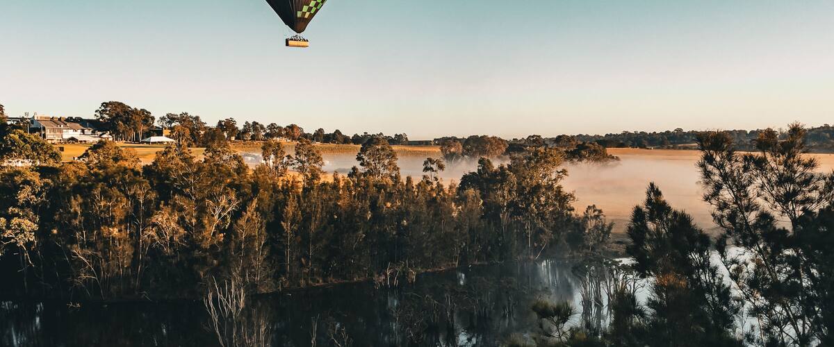 Un ballon à air chaud survole un lac paisible, dont la surface reflète les arbres et un paysage lointain.