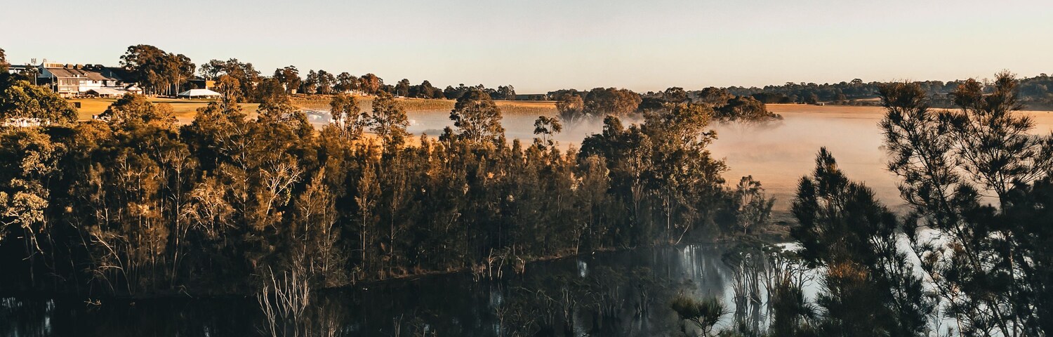 Um balão de ar quente voando sobre um lago calmo, com reflexos de árvores e uma paisagem distante.