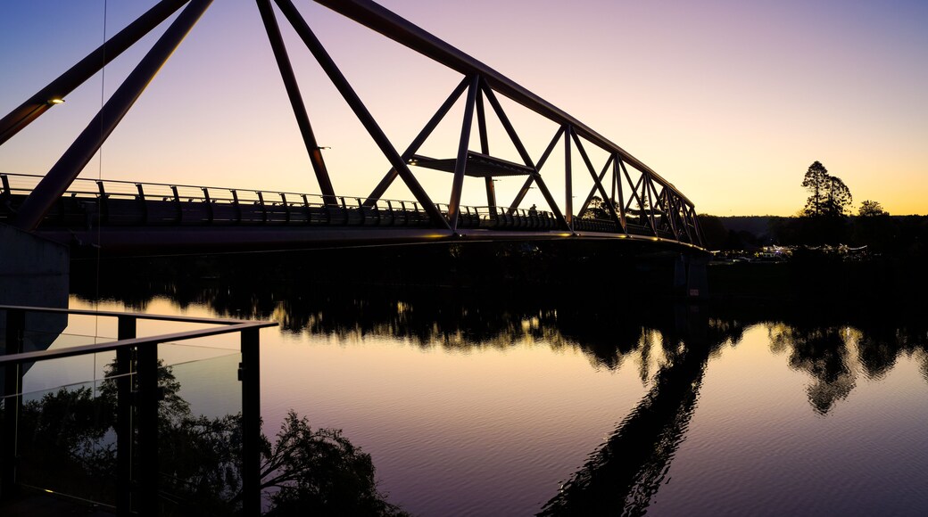A steel bridge with a triangular truss design spanning over a calm body of water, reflecting the structure and the sky.