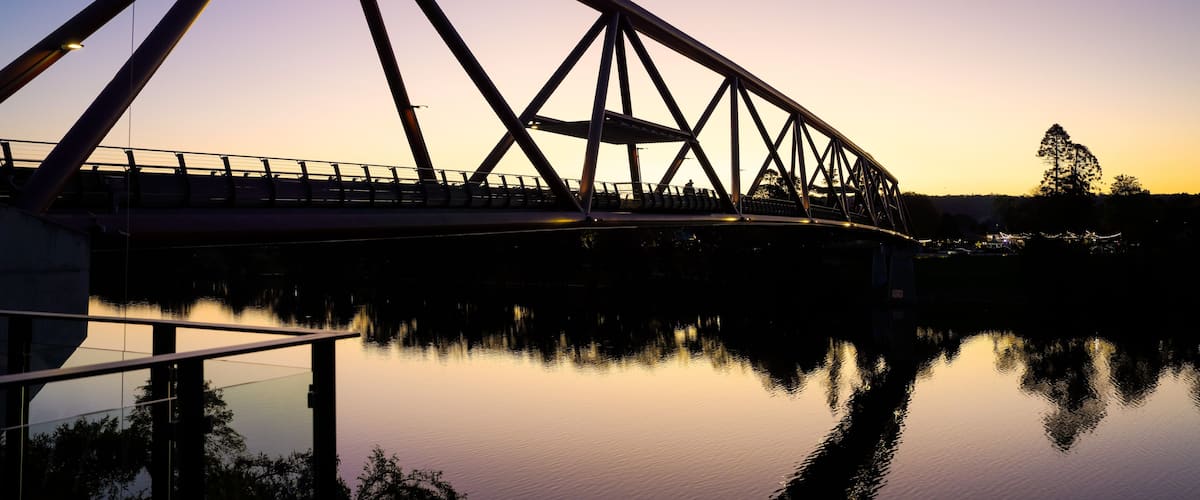 A steel bridge with a triangular truss design spanning over a calm body of water, reflecting the structure and the sky.