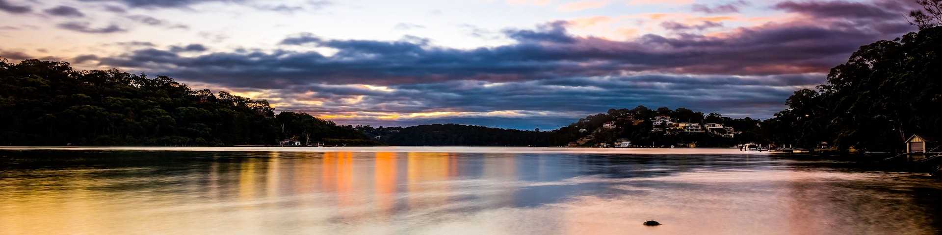 Um lago calmo sob um céu de pôr do sol, cercado por árvores e uma linha de costa distante com edifícios.