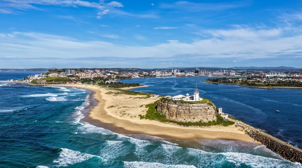 A coastal cityscape with a prominent white lighthouse on a rocky outcrop, a sandy beach, and a clear blue sky.