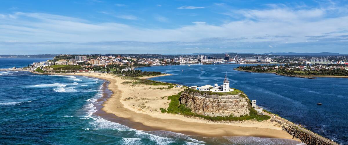 A coastal cityscape with a prominent white lighthouse on a rocky outcrop, a sandy beach, and a clear blue sky.