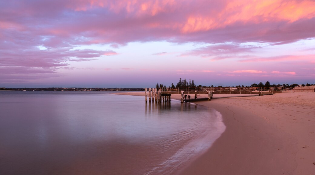A beach with a wooden pier extending into calm waters, surrounded by a sandy shore and a backdrop of a colorful sky.