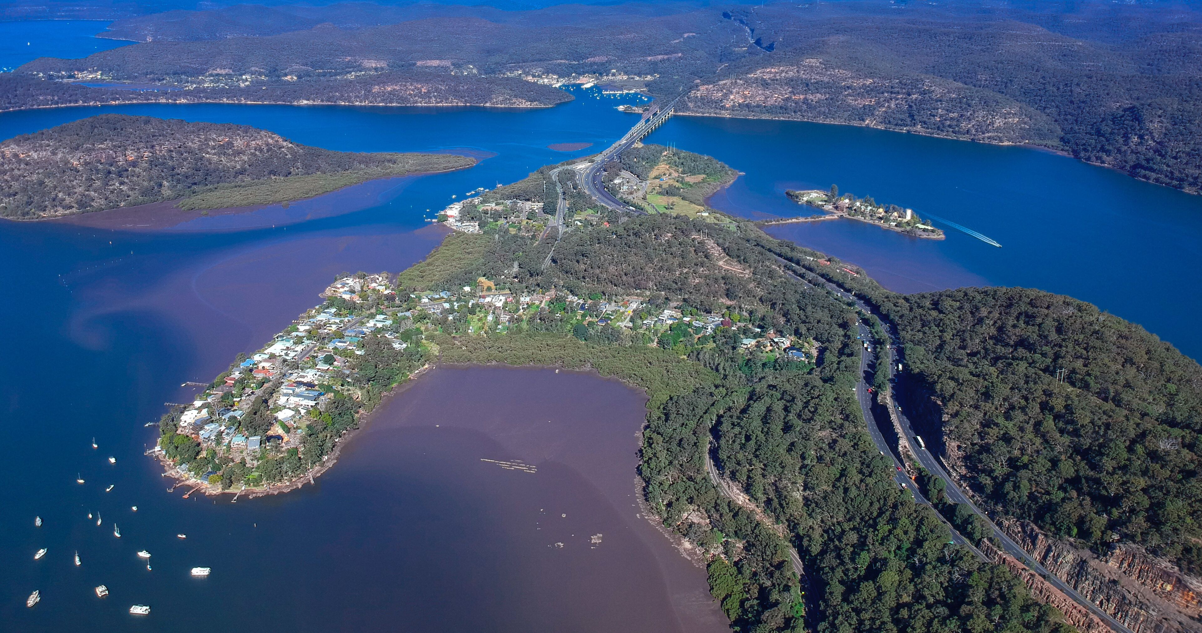 An aerial view of a coastal town with a bridge connecting two landmasses, surrounded by water and forested hills.