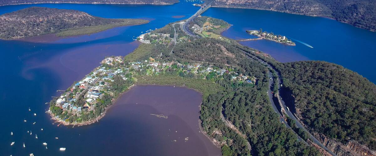 An aerial view of a coastal town with a bridge connecting two landmasses, surrounded by water and forested hills.