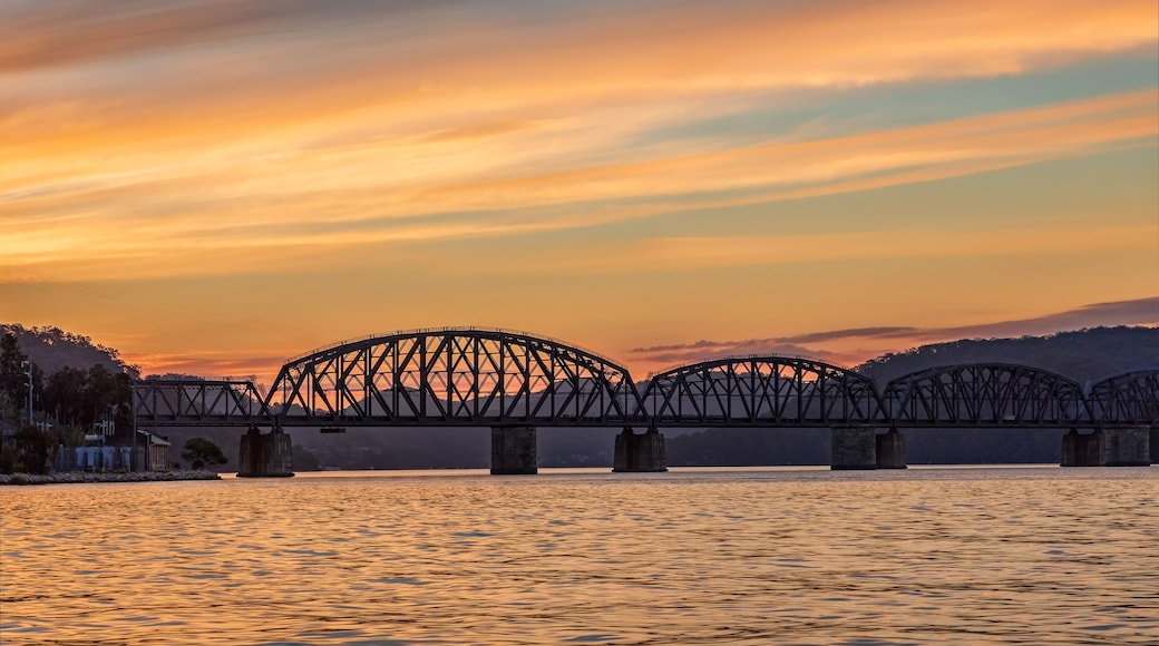 A steel bridge spanning a river at sunset with a mountainous backdrop.